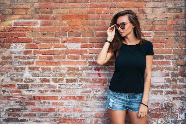 outdoor portrait of a young pretty hipster woman with long dark hair wearing black blank t-shirt and blue jeans shorts standing against brick wall background