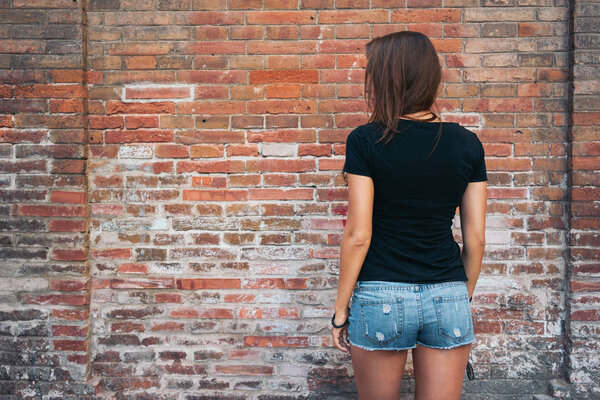 outdoor portrait of a young pretty hipster woman with long dark hair wearing black blank t-shirt and blue jeans shorts standing against brick wall background