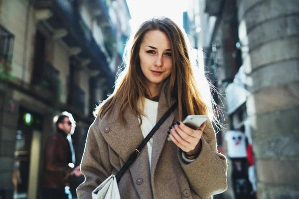 Half length portrait of young woman holding a mobile phone in her hand looking at the camera on blurred city street background