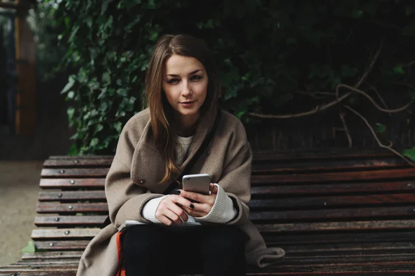 Молодая Женщина Модном Наряде Используя Смартфон Сидя Деревянной Скамейке Открытом — стоковое фото