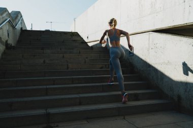 Genç, sportif bir kadın dışarıda antrenman yaparken enerji dolu bir şekilde su şişesiyle yukarı koşuyor. Koşucu kadın iyi durumda. Sabah koşusunu bitiriyor. Merdivenleri koşarak çıkıyor..