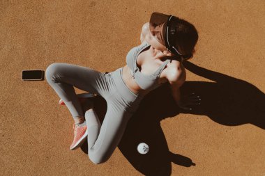 Spor kıyafetleri giyen beyaz çekici bir kadın dışarıda antreman yaptıktan sonra gün ışığının tadını çıkarıyor. Spor salonunda elinde su şişesi ve akıllı telefonuyla dinlenen bir kız.