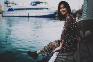Çekici, çekici Asyalı model uzun siyah saçlı, maksimum şifonlu elbise giyen kadın, şehrin rıhtım bölgesinde bulanık bir yat arka planında otururken kameraya mutlu bir şekilde gülümsüyor..