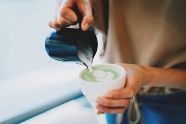 Barista lezzetli ve sağlıklı sütlü sütlü çay yaparken şablon yapıyor. Bir kadının eli, modern bir Asya kahve dükkanında taze yapılmış doğal eşleşen çay ile sıcak kremşantili sütü fincana döküyor..