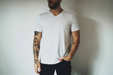 Sakallı ve dövmeli genç hippi beyaz bir tişört giymiş, beyaz bir duvara karşı poz veriyor. Logo ya da tasarım için boş alan. Tişört maketi.