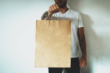 Beyaz tişört giyen genç hippi logo ya da tasarım için boş yeri olan el işi kâğıt paketleri tutuyor, saplı alışveriş çantasının maketi..