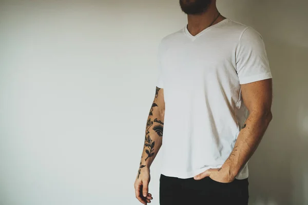 Male hipster with beard and tattoos wearing a blank white t-shirt and black jeans is standing on a white wall background. Horizontal mock up. Empty space for text o design.