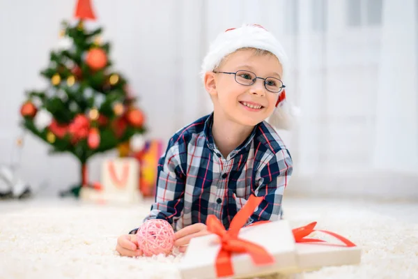 Niño con un sombrero rojo de Navidad tirado en el suelo y abre sus regalos de Navidad —  Fotos de Stock