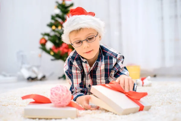 Niño con un sombrero rojo de Navidad tirado en el suelo y abre sus regalos de Navidad —  Fotos de Stock