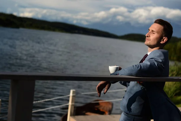 The groom in a suit on the waterfront drinks coffee