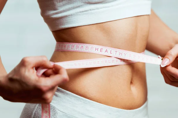 Beautiful, fit, young woman measuring her waist with a measuring tape, with word HEALTHY written on it, close up