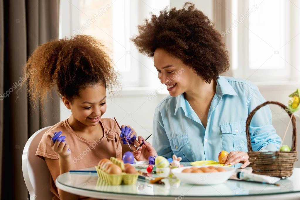 Beautiful African American woman and her daughter coloring Easter eggs at the table  
