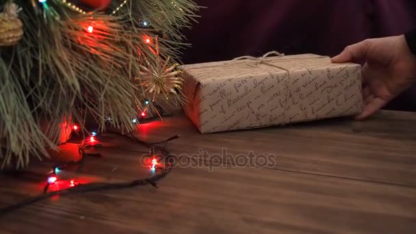 남자는 크리스마스 트리 아래 선물 상자를 넣습니다. 전나무 나무 테이블에 서 서와 빛나는 갈 랜드와 크리스마스 장난감 장식. — 비디오
