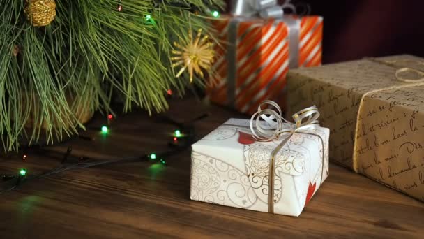 Коробки подарунків знаходяться під деревом. Подарунки упаковані в різнокольоровий папір і пов'язані нитками і стрічками. Різдвяна гірлянда на гілках ялинки. Гілки дерева крупним планом, пересування, панорама — стокове відео