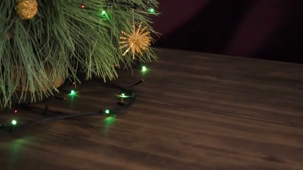 Quell'uomo mette una scatola di regali sotto l'albero di Natale. Cavalletto in abete sul tavolo di legno e decorato con ghirlanda incandescente e giocattoli di Natale . — Video Stock