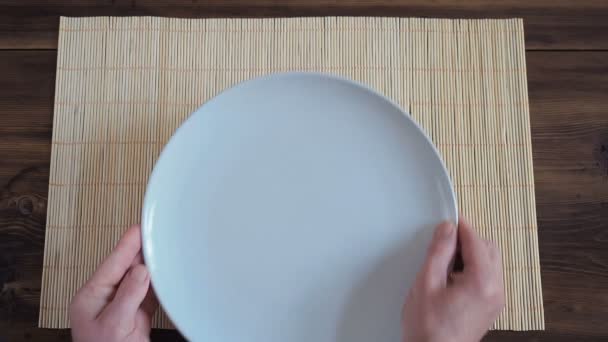 De man zet op houten tafel van de oude planken met servet vervaardigd van bamboe, een witte ronde schotel en een metalen vork — Stockvideo