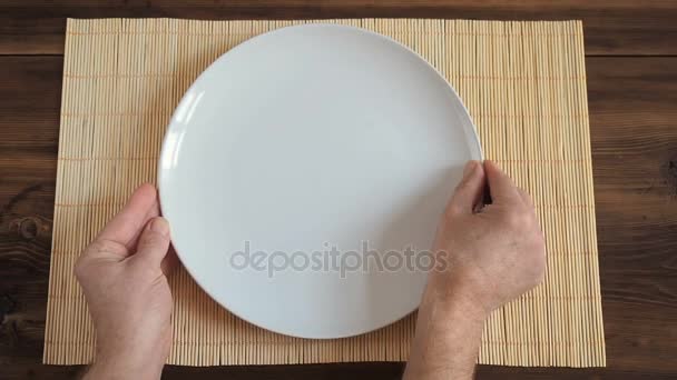 Muž klade na dřevěný stůl ze starých desek s ubrousek vyrobené z bambusu, bílý kulatý talíř a kovové vidlice a bílé papírové ubrousky — Stock video