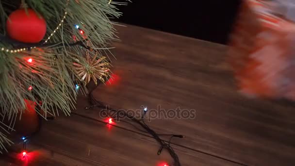 Człowiek stawia paczek z prezentami pod choinkę. Jodła stanąć na drewnianym stole i ozdobione świecącą garland i Boże Narodzenie zabawki. — Wideo stockowe