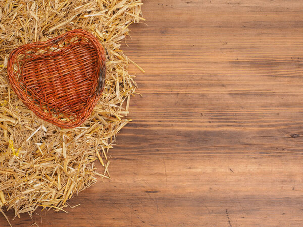 Rural eco background. Wicker-work basket from withe in heart shape with straw on the background of old wooden planks. The view from the top. Creative background for greeting cards