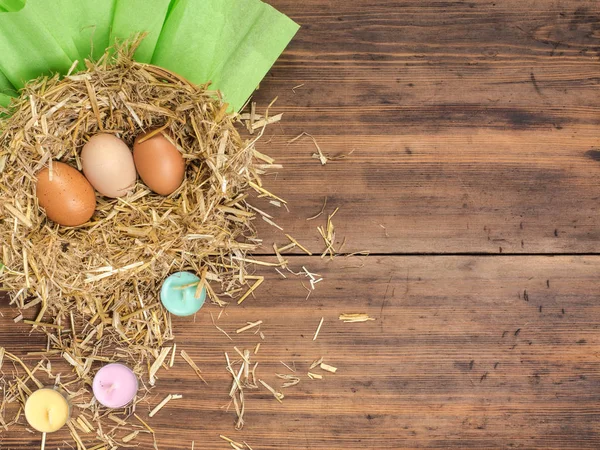 Kahverengi yumurta saman içinde iç içe. Kırsal çevre arka plan ile kahverengi tavuk yumurtası, saman, renkli mumlar ve eski ahşap plakalar arka plan üzerinde kağıt. Üstten Görünüm. Paskalya kartları veya menü yaratıcı arka plan