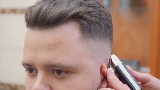 Stylist schneidet Männchen Schläfen mit Clipper, Nahaufnahme. Meister bei der Arbeit im Friseursalon. Mann im Friseurladen. selektiver weicher Fokus. verschwommener Hintergrund. — Stockvideo