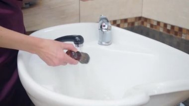 Kuaför fırçayla tıraş köpüğü hazırlıyor, yakın çekim. Beyaz lavabo ve bej kaplamalı fayans, berber dükkanının içi. Seçici yumuşak odaklanma. Bulanık arkaplan.