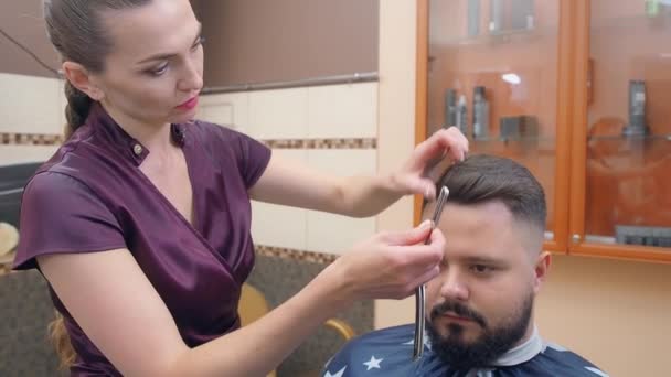 Female in dark red dress cutting mans hair with open razor, medium shot. Male in hairdressing saloon. Interior of barbershop. Selective soft focus. Blurred background.