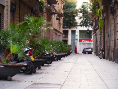 Barselona, İspanya - Ağustos 2019: Yeşil bitkiler arasında motosiklet parkı, orta manzara. Binalar arasındaki asma kat. Sokakta yürüyen bir kadın, araba süren. Seçici yumuşak odaklanma. Bulanık arkaplan.