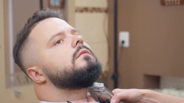 Мужчина получает услуги в парикмахерской, с близкого расстояния. Стилист стрижет бороду мужчинам клиппером и черной расческой. Интерьер парикмахерской. Селективный мягкий фокус. Размытый фон . — стоковое видео