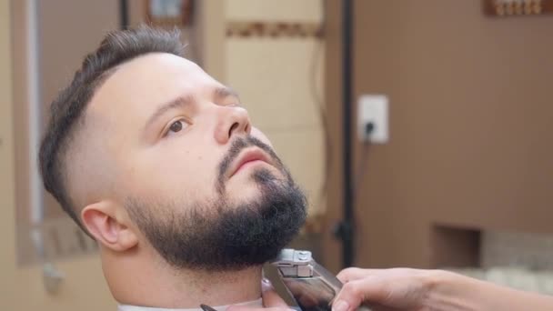 Hombre recibiendo servicio en la barbería, tiro cerrado. Estilista cortando barba de hombre con clipper y peine negro. Interior del salón de peluquería. Enfoque suave selectivo. Fondo borroso . — Vídeo de stock