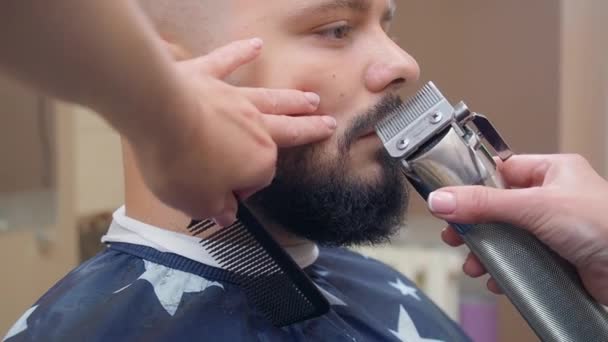 Man i frisersalong, närbild. Stylist skära mans skägg med clipper och svart kam. Inspelad i frisörsalongen. Selektivt mjukt fokus. suddig bakgrund. — Stockvideo