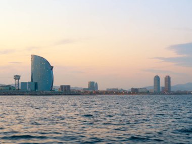 Barselona, İspanya - Ağustos 2019: deniz yüzeyi dalgalanması. Deniz kenarındaki modern yüksek binalar, ekstra uzun manzaralı. Ufuktaki sisli tepeler. Bulutlu gökyüzü. Seçici yumuşak odaklanma. Bulanık arkaplan.