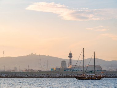 Barselona, İspanya - Ağustos 2019: Turistlerle dolu bir yat kıyı boyunca seyrediyor, ekstra uzun bir manzara. Deniz kenarındaki modern binalar. Ufuktaki sisli tepeler. Bulutlu gökyüzü. Seçici yumuşak odaklanma. Bulanık arkaplan.