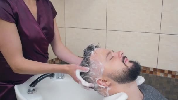 Master in dark red dress washing clients head with shampoo in white washstand, close shot. Male in hairdressing saloon. Selective soft focus. Blurred background. — Stock Video