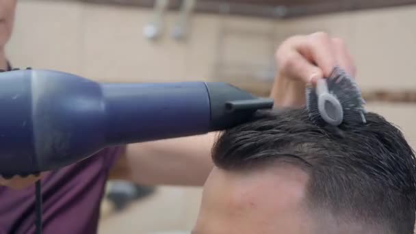 Stylist making modern hairdo with hair dryer and round comb, close shot. Male in hairdressing saloon. Selective soft focus. Blurred background. — 비디오