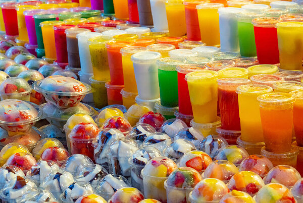 Multicolour background of fresh smoothie and fruits cutted into pieces in plastic cups, close up view. Selective soft focus. Blurred background.