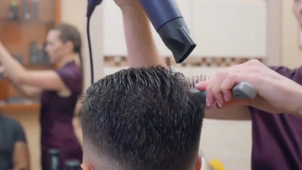 Female in dark red dress drying clients hair with dryer and round comb, close shot. Male in hairdressing saloon. Interior of barbershop. Selective soft focus. Blurred background. — Stock Video