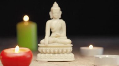The movement to the statue of Buddha from blur to blur. White figure of buddha sitting in a meditation pose surrounded by burning candles. Slowly sliding, close-up shot from defocus