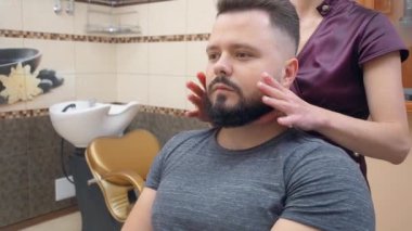 Barber moistening males beard with mousse, medium shot. Male in barbershop. Interior of hairdressing saloon. Selective soft focus. Blurred background.