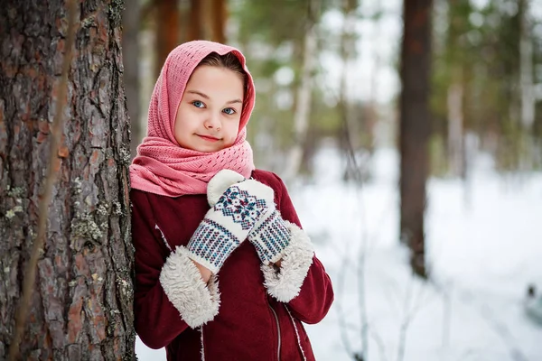 美丽的女孩在一个粉红色的头围巾和针织手套站在树在冬季森林 — 图库照片