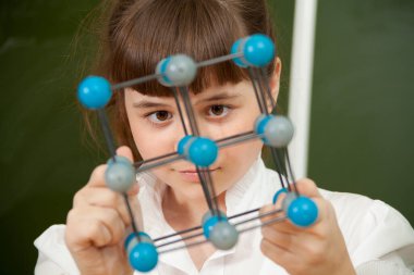 Tahtanın yanındaki kız, moleküllerin modelini elinde tutuyor.