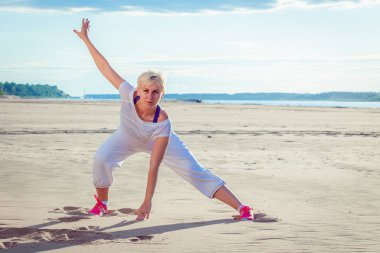 Kumsalda beyaz eşofmanlı bir kadın spor yapıyor.