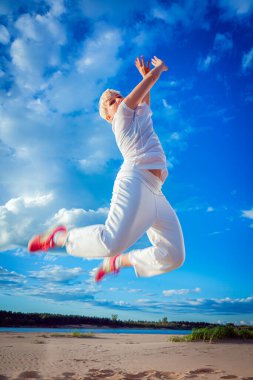 Beyaz eşofmanlı bir kadın kumlu bir plajda mavi gökyüzüne karşı zıpladı.