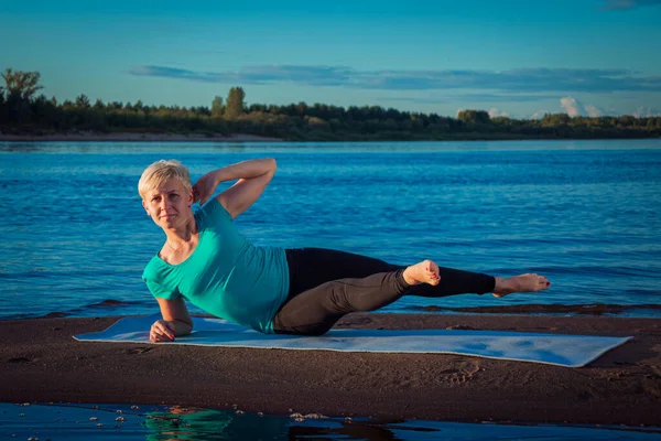 blonde woman with a short haircut on a sandy beach near the water lies on a blue rug and does exercises for the abs