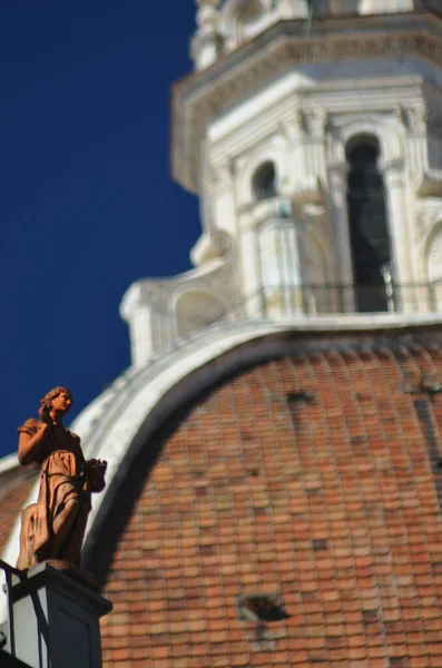 Küçük bir terracotta melek heykeli, elini kaldırmış, bir çatının köşesinde. Floransa 'nın Duomo kubbesinin arkasında (ve odak dışında) yükselir. Gökyüzü mavi.
