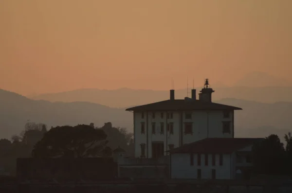The sun is setting over the hills outside of Florence, turning the sky shades of orange. Cypress trees are seen in silhouette. Vapour trails are in the sky.