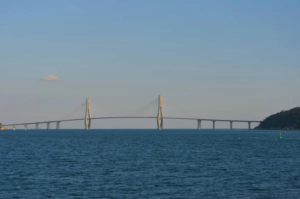 A bridge arches over a bay from an island  to the mainland. Two pylons, with radiating supportive metal struts, rise above the bridge to form an architectural feature. The sky is blue with clouds.