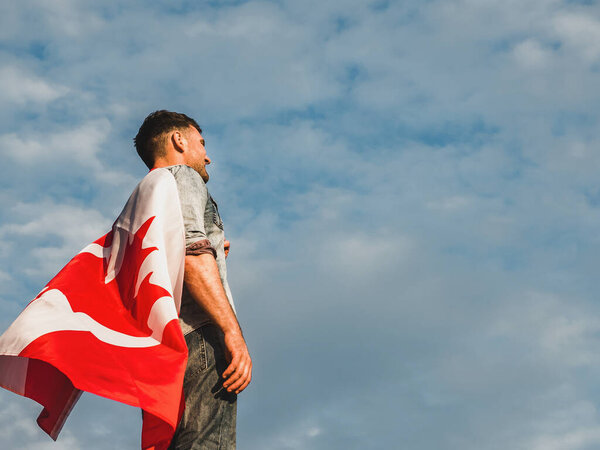 Attractive man holding Canadian Flag on blue sky background on a clear, sunny day. View from the back, close-up. National holiday concept