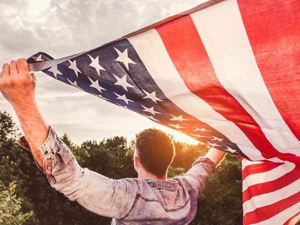 Attractive man holding Flag of the United States on blue sky background on a clear, sunny day. View from the back, close-up. National holiday concept