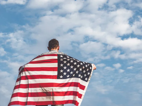 Attractive man holding Flag of the United States on blue sky background on a clear, sunny day. View from the back, close-up. National holiday concept